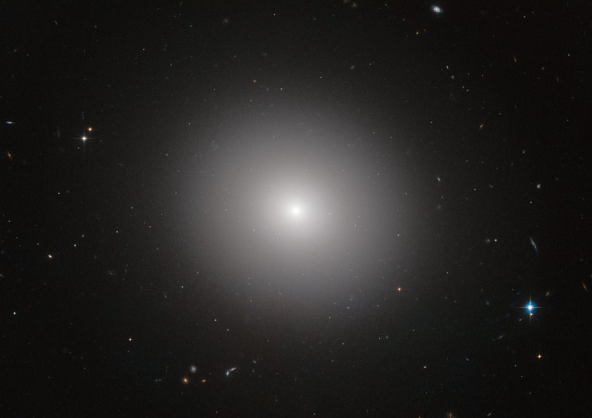 An image of an elliptical galaxy known as IC 2006 taken by the Hubble Space Telescope.  It appears as a bright, roughly round mass of white stars with density increasing steadily towards the center.  There are many other small stars and galaxies visible in the background. Public domain.